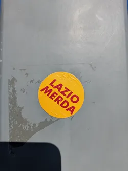 Street sticker This is a round, yellow sticker with the words "LAZIO MERDA" printed in red. The sticker is stuck on a gray surface, possibly a wall or door. The text is in bold and slightly tilted.