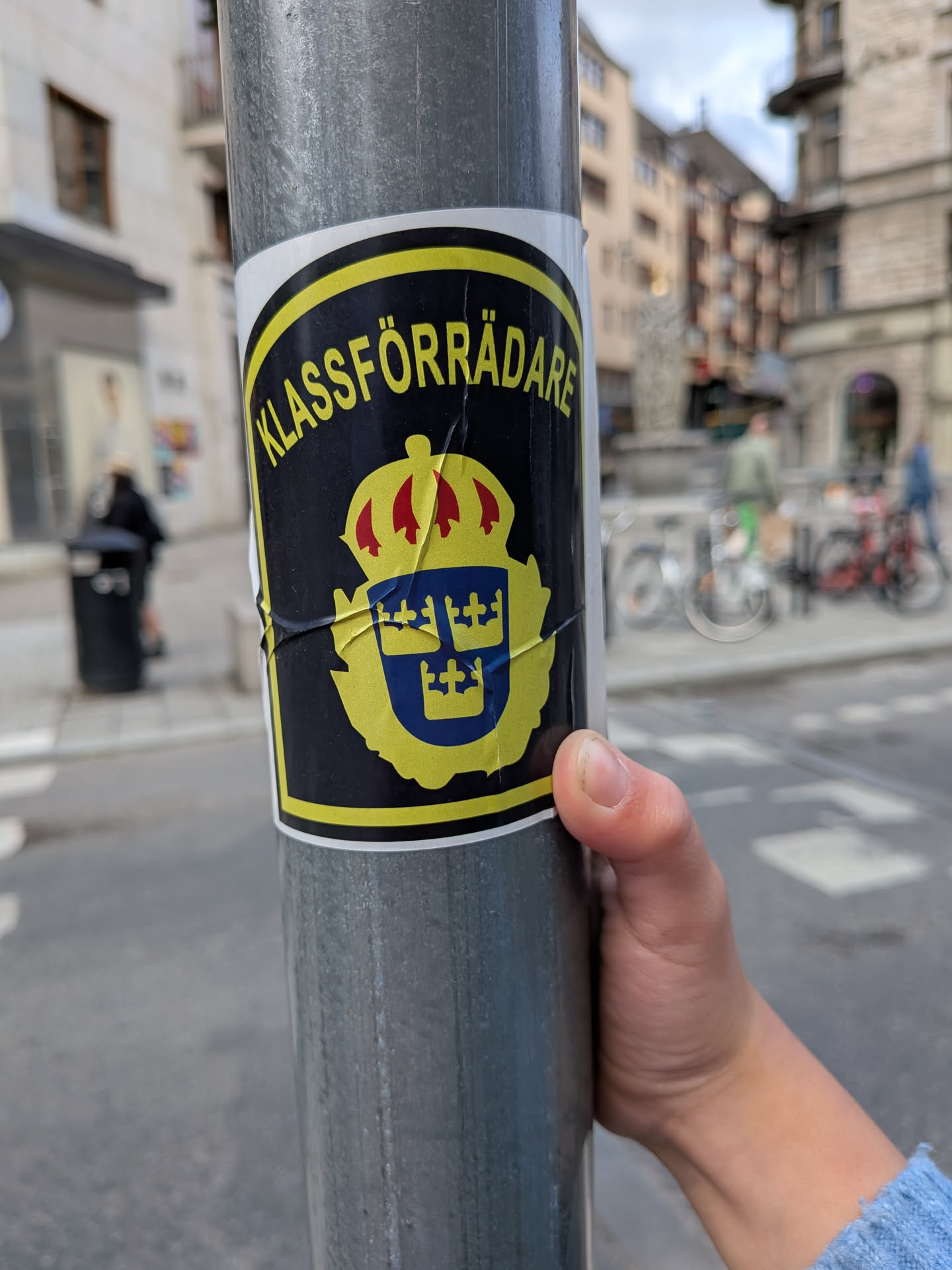 A shield-shaped sticker on a metal pole. It features a black background, yellow border, and the Swedish text 'KLASSFÖRRÄDARE' arched over a parody of the Swedish police coat of arms with the three crowns.