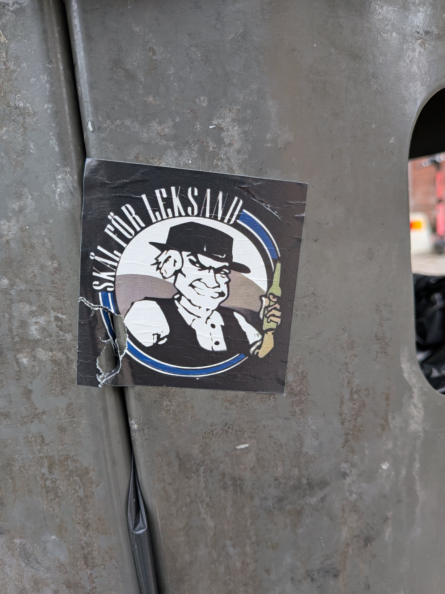 Square black sticker featuring a circular graphic of a cartoon man in a hat holding a bottle. Includes the Swedish text 'SKÅL FÖR LEKSAND' in white, bordered by blue and white stripes.