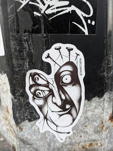 Street sticker A black and white sticker depicting a surreal image. A human face appears to be merging with a butterfly, with several nails piercing the top of the head. The style is reminiscent of dark art or surrealism.  The sticker has the Instagram handle '@ig.bartolo.tattoo' printed on it.