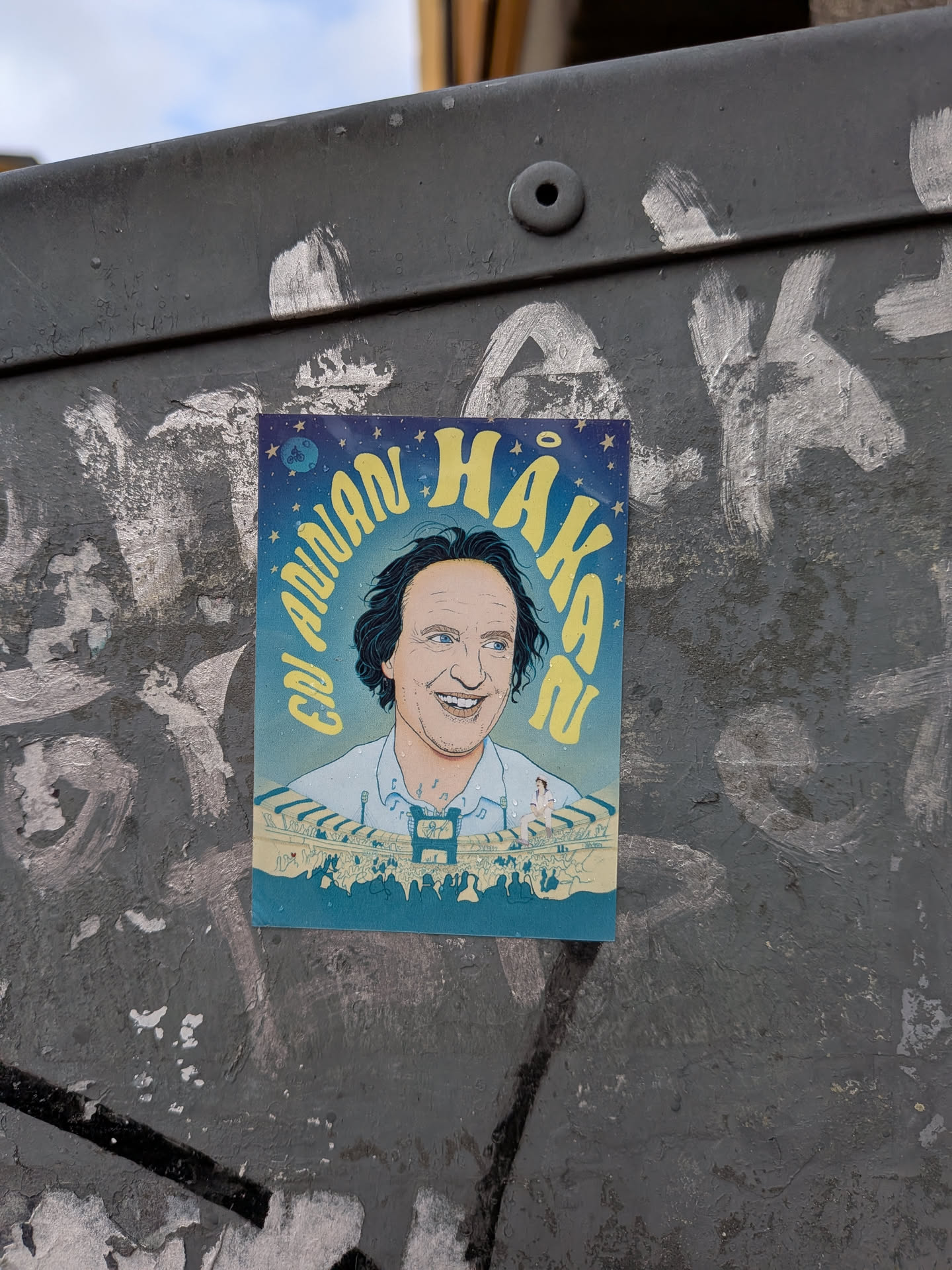 The sticker features a vibrant, stylized illustration of a smiling man with dark, wavy hair and blue eyes, wearing a light blue collared shirt. He is depicted from the chest up, looking towards the right with a wide, joyful smile. The background is a deep blue night sky adorned with numerous yellow stars of varying sizes. Curving around the man's head, in bright yellow, stylized letters, is the text "EN ANNAN HÅKAN". Below the man, a lively concert scene unfolds with a stage, bright spotlights, and a dense crowd of silhouetted figures. A small, whimsical detail of a person riding a bicycle on the moon is visible in the top-left corner. The overall aesthetic is cheerful, colorful, and reminiscent of a concert poster or pop art.