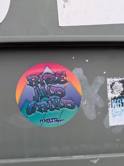 Street sticker A round sticker with the words "Rise and Grind" in stylized graffiti font, set against a mountain range illustration. The sticker has a vibrant color gradient, with pink and orange at the top fading to blue and green at the bottom. The URL stickercosmos.com is printed at the bottom.