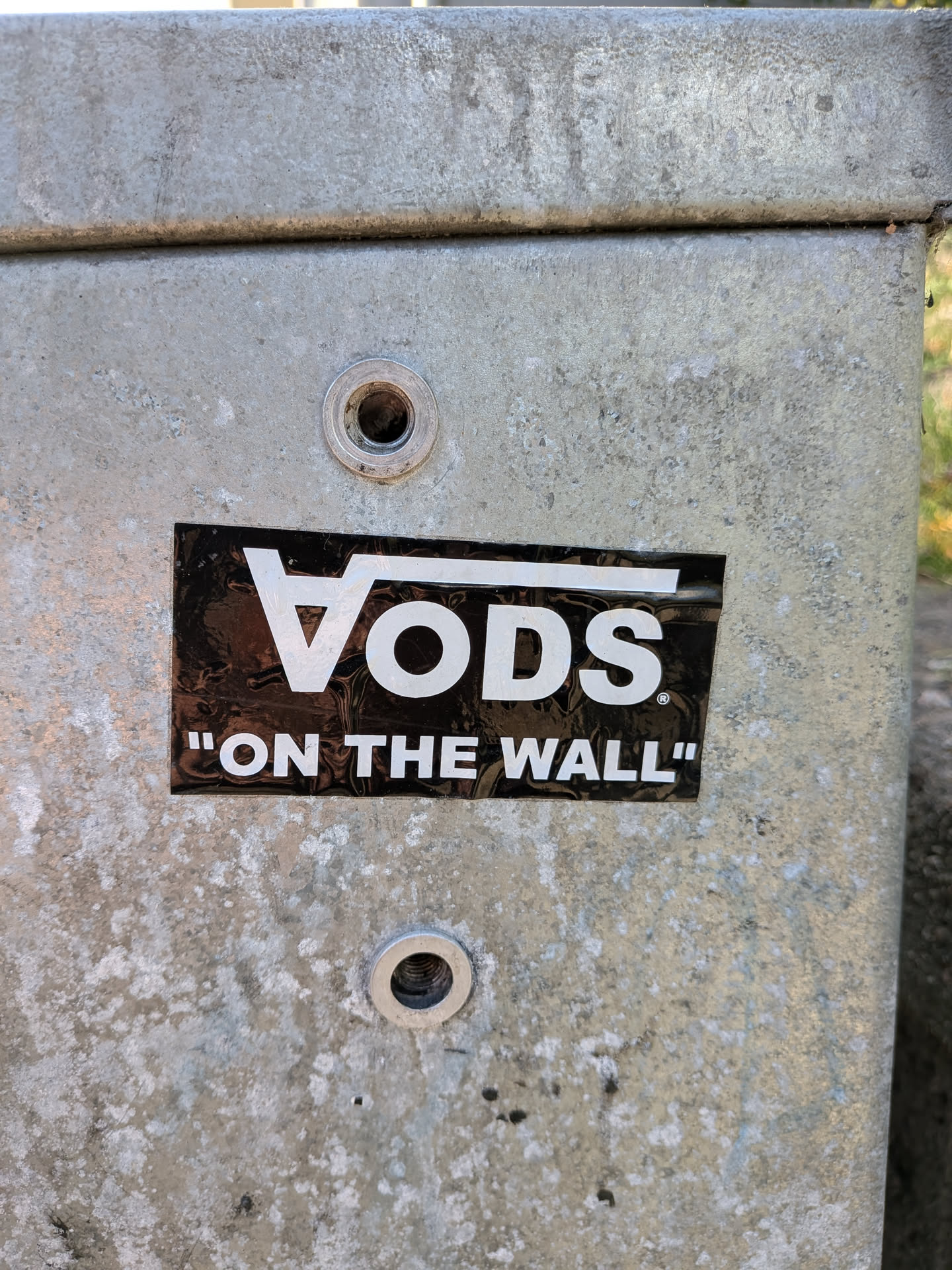 The sticker is rectangular with a black glossy background. The main element is the Vans logo, stylized as 'VO DS', with the 'V' appearing as a triangle and the 'O' as a circle. The letters are in a bold white font. Below this, in smaller white text enclosed in quotation marks, is the phrase 'ON THE WALL'. The sticker shows some signs of wear and tear, with minor scuffs and reflections on its surface, indicating it is a genuine sticker applied to a metal surface.