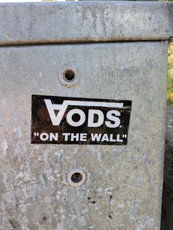 Street sticker The sticker is rectangular with a black glossy background. The main element is the Vans logo, stylized as 'VO DS', with the 'V' appearing as a triangle and the 'O' as a circle. The letters are in a bold white font. Below this, in smaller white text enclosed in quotation marks, is the phrase 'ON THE WALL'. The sticker shows some signs of wear and tear, with minor scuffs and reflections on its surface, indicating it is a genuine sticker applied to a metal surface.