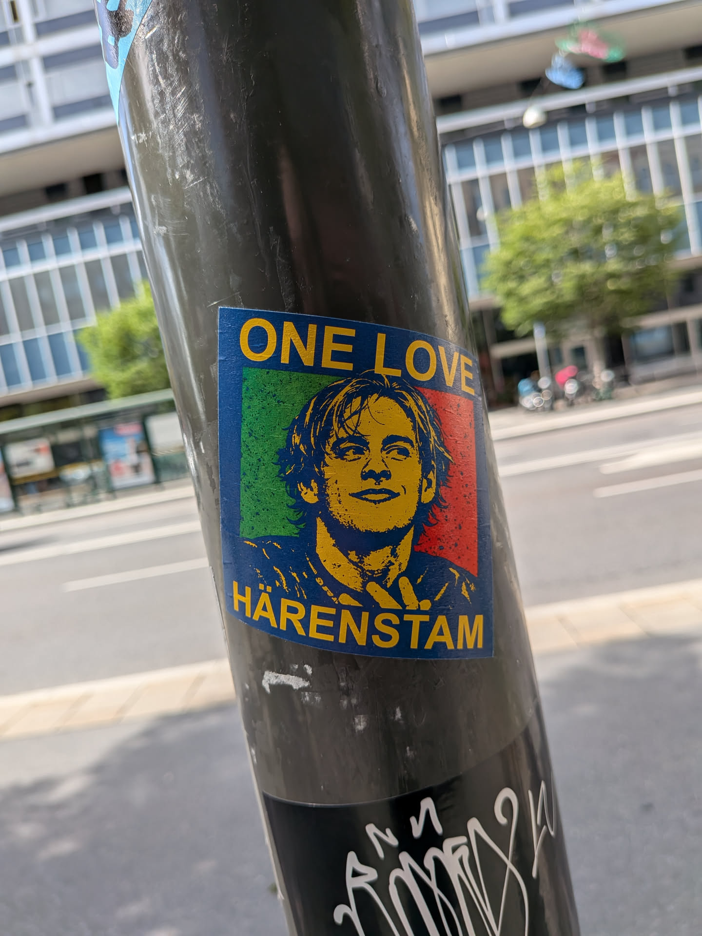 A rectangular sticker on a pole showing a yellow stencil portrait of Magnus Härenstam. The background is split green and red, framed in blue, with yellow text 'ONE LOVE' above and 'HÄRENSTAM' below.