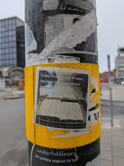 The sticker is square-shaped and features a printed image of an open book with text overlaid. The text is in a light color against a darker background, making it somewhat difficult to read. The image of the book appears to be a stylized illustration or photograph. The sticker is affixed to a pole that is wrapped in reflective yellow tape. The overall appearance suggests a religious or inspirational message.