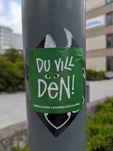 A square, slightly crumpled green sticker attached to a gray pole. The sticker features the text "Du Vill Den!" in bold white letters, with two small circles above the text that resemble eyes. Below the main text, smaller text reads "GRÖNA LINJEN • BIOPREMIÄR 25 APRIL".  The sticker is adhered to a gray pole, partly covering another black and white sticker with a diamond and curved line design.