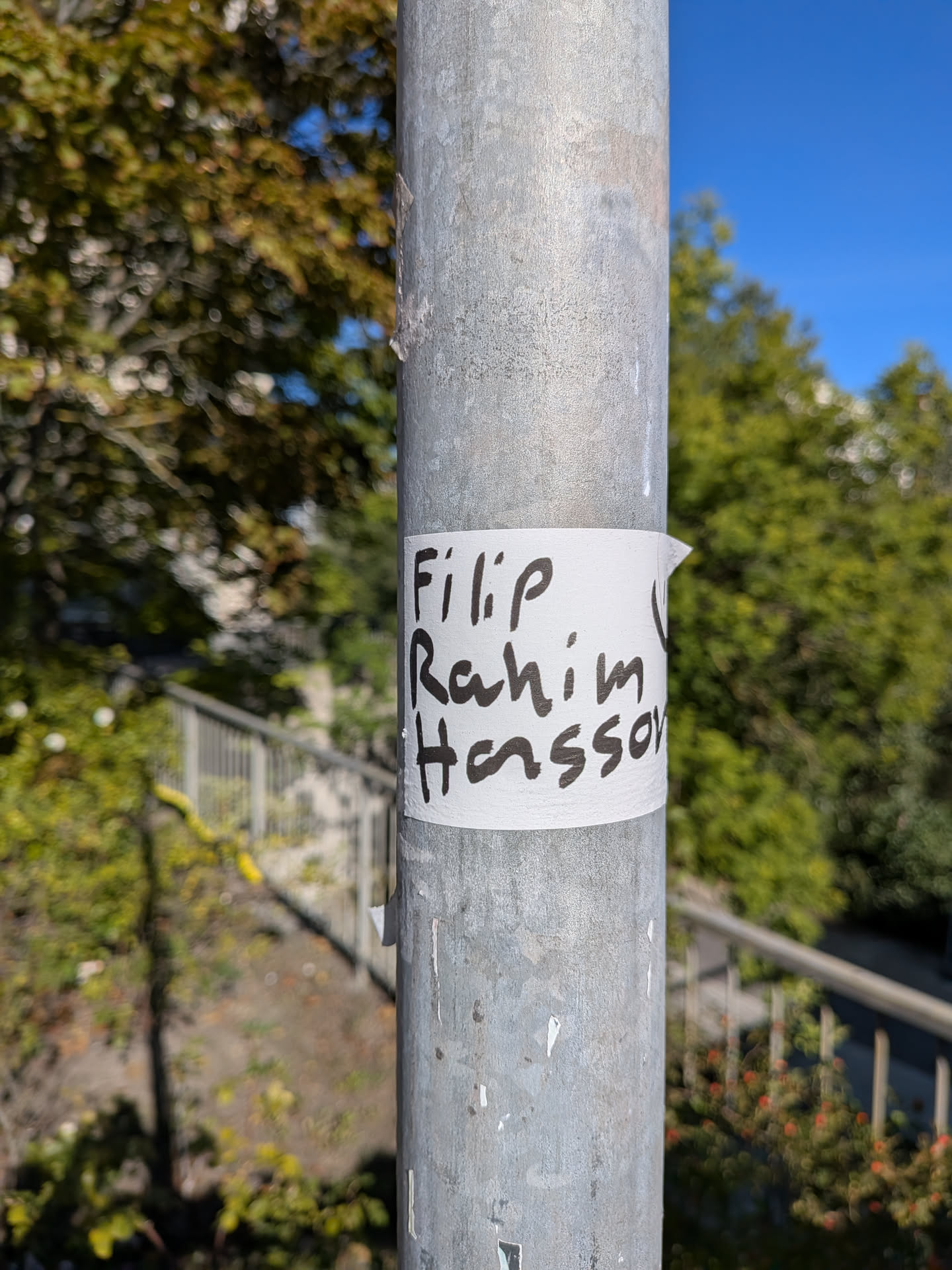 The sticker is a piece of white paper wrapped around a metal pole. It has handwritten text in black marker. The text appears to be a name or names, written in a casual, slightly messy script. The edges of the sticker are somewhat rough, and it is attached with what looks like tape on the right side, which is slightly peeled back. The background is out of focus, showing greenery and a blue sky.