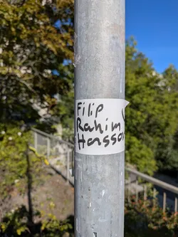 Street sticker The sticker is a piece of white paper wrapped around a metal pole. It has handwritten text in black marker. The text appears to be a name or names, written in a casual, slightly messy script. The edges of the sticker are somewhat rough, and it is attached with what looks like tape on the right side, which is slightly peeled back. The background is out of focus, showing greenery and a blue sky.