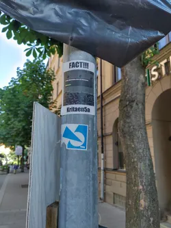 Street sticker A sticker featuring an image and the text 'FACT!!!' and 'Kritae5a' is affixed to a metal pole. The image appears to show a crowd of people.  The sticker's style suggests a meme or internet-based origin. There's another blue and white sticker below it, but less visible.