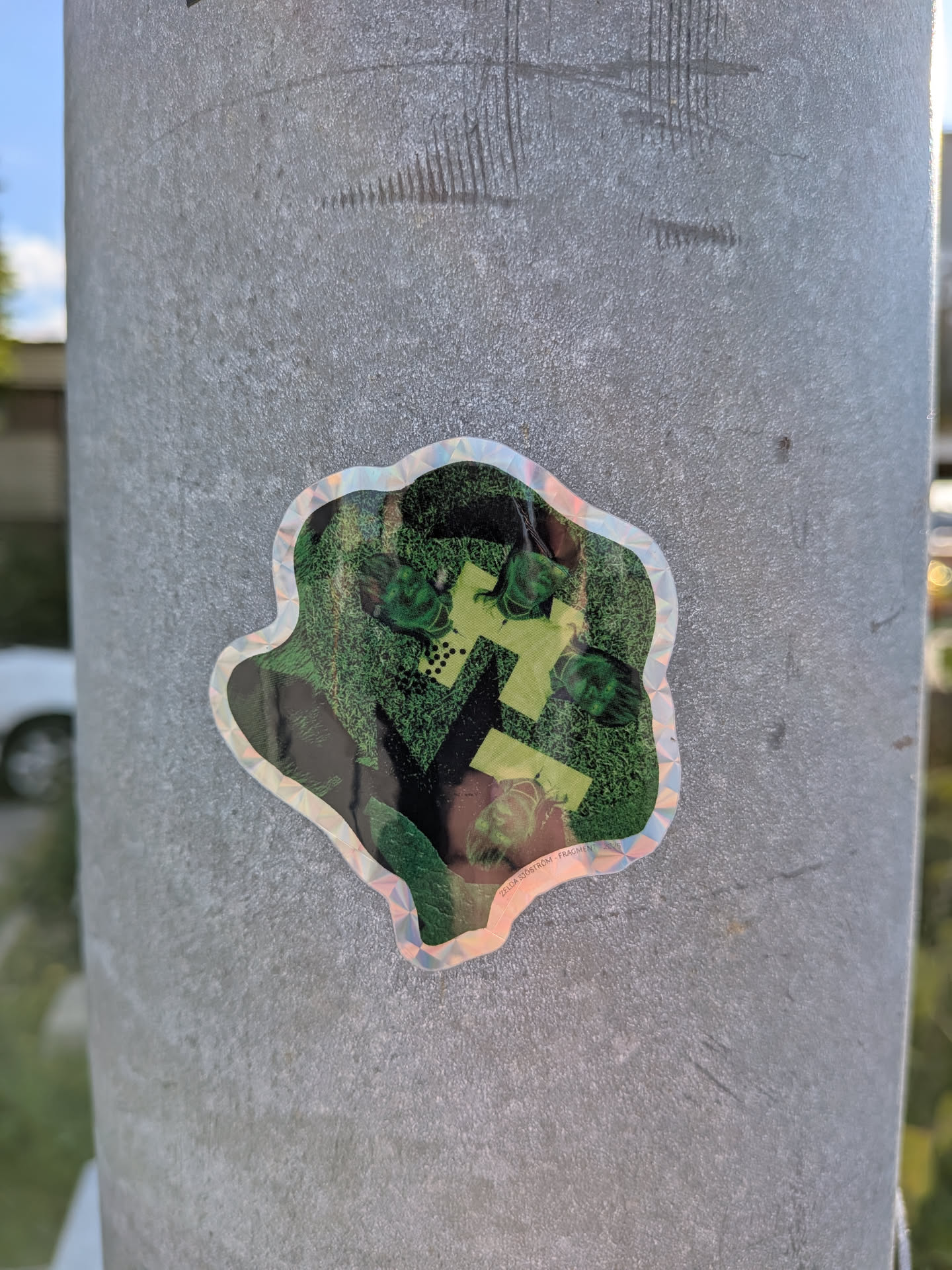 The sticker has an irregular, wavy border with a holographic sheen that reflects light in rainbow colors. The central design appears to be a stylized representation of the Triforce symbol, composed of green triangular shapes and yellow segmented lines, set against a dark background with hints of green foliage or grass texture. Several small, green, creature-like or stylized head elements are visible within the design, potentially representing characters or items from a video game. There is also a small line of text at the bottom edge of the sticker.