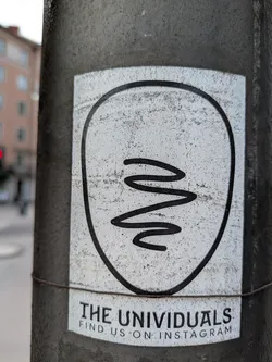 Street sticker This sticker on a pole features a minimalist design with a squiggly line inside a rounded frame. Below the design, the text 'THE UNIVIDUALS' is printed, followed by 'FIND US ON INSTAGRAM', indicating a social media presence.