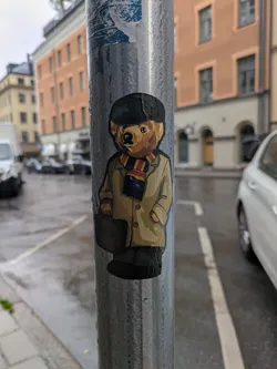 Street sticker A sticker of a bear dressed in a cap, coat, and scarf, holding a briefcase, is affixed to a metal pole. The bear gives off a professional and sophisticated vibe.