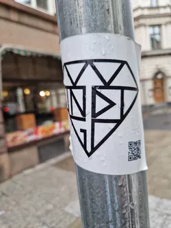 Street sticker A white sticker with black graphic design.  The design is a diamond shape incorporating an arrow pointing upwards and the letters INDIG. A QR code is present on the bottom right corner of the sticker. The sticker is affixed to a weathered metal pole. 