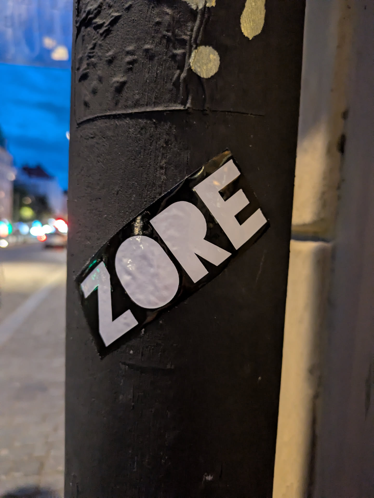 The sticker is a rectangular, horizontally oriented design featuring the word 'ZORE' in bold, white, sans-serif capital letters set against a glossy black background. The 'O' character is particularly prominent due to its round, solid white fill. The sticker appears to have a slightly crinkled or uneven surface, especially visible within the white letters due to light reflection, suggesting it's adhered to a textured surface or has seen some wear. It is affixed diagonally to a dark, textured pole.