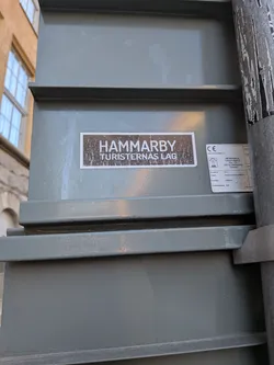 Street sticker A rectangular sticker with a distinct white border, featuring a dark brown or black background with a distressed, grunge-like texture. White, sans-serif text is prominently displayed in two lines: 'HAMMARBY' on the top, and 'TURISTERNAS LAG' below. The sticker is affixed to a grey-green metal utility box or structure.