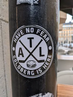 Street sticker The sticker is circular with a prominent black outer ring and a white inner circle. The outer ring contains the text 'HAVE NO FEAR' arching across the top and 'GOLDBERG IS HERE' arching across the bottom, all in white capital letters. The inner white circle features two black hockey sticks crossed in an 'X' shape. Within the quadrants formed by the sticks, there are black capital letters: 'A' in the lower left, 'T' in the upper center, and 'L' in the lower right, forming 'ATL'. Centered at the bottom of the inner circle, below the crossed sticks, is a black outline drawing of a hamburger with sesame seeds on the bun. The design is entirely monochrome, black and white, and shows minor scuffs and wear consistent with being a physical sticker on a pole.