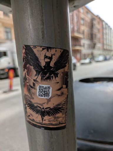Street sticker The sticker features a stylized image of a figure with wings, wearing a mask. The QR code might lead to more information about the artist or the image itself. The background is a faded tan, giving it a vintage or distressed look. There is a bird below the masked figure.