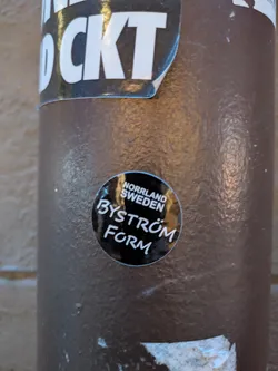 Street sticker The sticker is circular and predominantly black with white text. The top of the circle contains the text 'NORRLAND SWEDEN' in smaller, capitalized letters. The main text in the center reads 'BYSTRÖM FORM' in a larger, stylized white font with a slight script-like appearance. The sticker has a glossy finish and is applied to a textured, brown cylindrical surface.