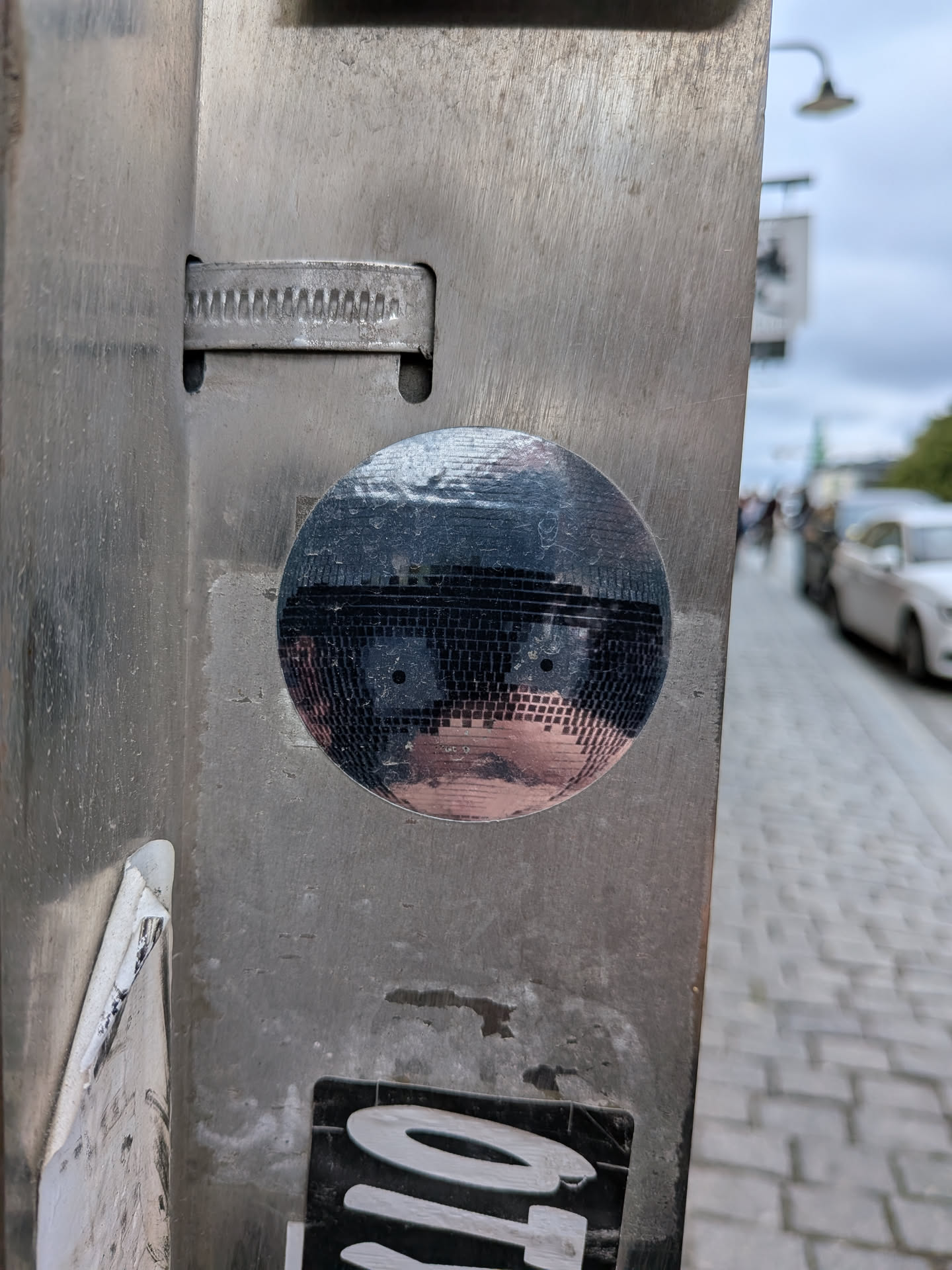 Street sticker The sticker is circular and predominantly black, mimicking the reflective surface of a disco ball. It features a stylized face with two dark, circular eyes and a pinkish-tan area that suggests a nose and cheeks, all set against a background of small, square, reflective facets. The overall effect is that of a somewhat surreal, geometric portrait.