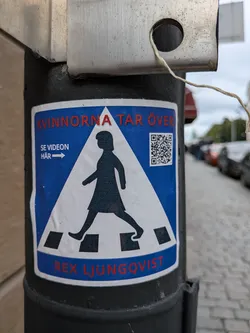 Street sticker The sticker is predominantly blue and white, designed to resemble a pedestrian crossing sign. At the top, in red capital letters, it reads "KVINNORNA TAR ÖVER". Below this, on the left side, there is white text "SE VIDEON HÄR" with a white arrow pointing to the right. A prominent black silhouette of a woman walking across a crosswalk is centered on the sticker. To the right of the silhouette, a QR code is visible. The bottom of the sticker features the name "REX LJUNGQVIST" in red capital letters on a blue band. The sticker appears slightly worn and adhered to a dark, cylindrical surface.