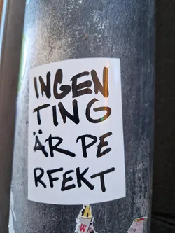 A white sticker with black hand-painted text on a gray pole. The text says "INGENTING ÄR PERFEKT", which translates from Swedish to English as "NOTHING IS PERFECT".