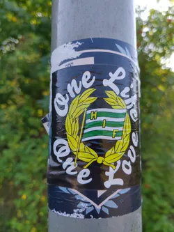 A black sticker with the text "One Life One Love" written in a stylized font.  At the center is a green and white flag with the letters "HIF" inside a yellow laurel wreath. The sticker is affixed to a grey pole, partially damaged along the edges. 
