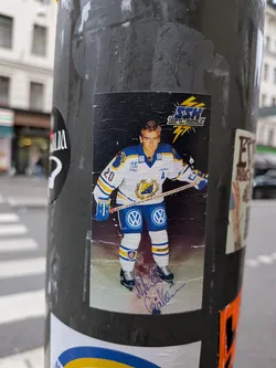Street sticker The sticker features a full-body image of a male ice hockey player in uniform, standing on an ice rink. He is wearing a white jersey with blue and yellow accents, blue pants with yellow stripes, and white skates. The jersey has the number '20' on the left sleeve and 'A' on the chest, along with several logos including 'Hyundai bilar', 'Jofa', and 'VW'. The player is holding a hockey stick. Above the player is a logo with 'SSH LIGHTNINGS' in stylized text with lightning bolts. The bottom of the sticker has a blue and white signature. The background is black, suggesting a studio or promotional photo. The sticker appears slightly worn and creased.
