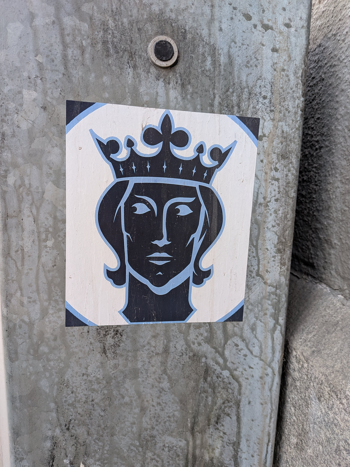 The sticker features a stylized portrait of a female figure wearing a crown. The main image is rendered in solid black, depicting the face and hair, with a prominent crown adorned with small diamond shapes. The eyes are open, looking slightly to the left, and the lips are closed. The black figure is outlined with a light blue border, which also defines the inner details of the eyes and the crown's embellishments. This central design is set against a white square background, which itself has light blue arcs in each corner, suggesting a circular motif within the square. The overall style is graphic and iconic.