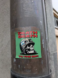 Street sticker The sticker is rectangular with a bright green background. Prominently displayed at the top are the words "DEATH DEATH" in bold, red, block letters with a slight shadow effect. Below this text is a detailed illustration of a human skull wearing a military-style helmet. The helmet is depicted in shades of light grey and black, with a blue Star of David emblem on its side. A red drip, resembling blood, extends from the helmet near the Star of David. The skull itself is rendered with intricate details, showing teeth and eye sockets. At the bottom of the sticker, also in bold red letters, is the phrase "TO THE IDF!". The overall aesthetic is graphic and impactful.