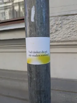 Street sticker A rectangular white sticker adhered to a grey metal pole. The sticker features black text in Swedish, centrally aligned. Below the text, there is a soft, irregular yellow-green gradient or spray paint effect across the bottom portion of the sticker. The sticker appears to be a printed label, showing slight wear and a hand-applied quality.