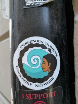 Street sticker The sticker is circular with a white background and a black, wavy border. In the center, a stylized indigenous woman with dark hair and brown skin is depicted in profile, looking to the right with her eyes closed. Her hair flows back and merges into a large, teal-colored spiral. The text 'INDIGENOUS WOMEN'S' curves around the top of the circle, and 'LEADERSHIP NETWORK' curves around the bottom. Below this main sticker, a portion of another black sticker with red text 'I SUPPORT' is visible.