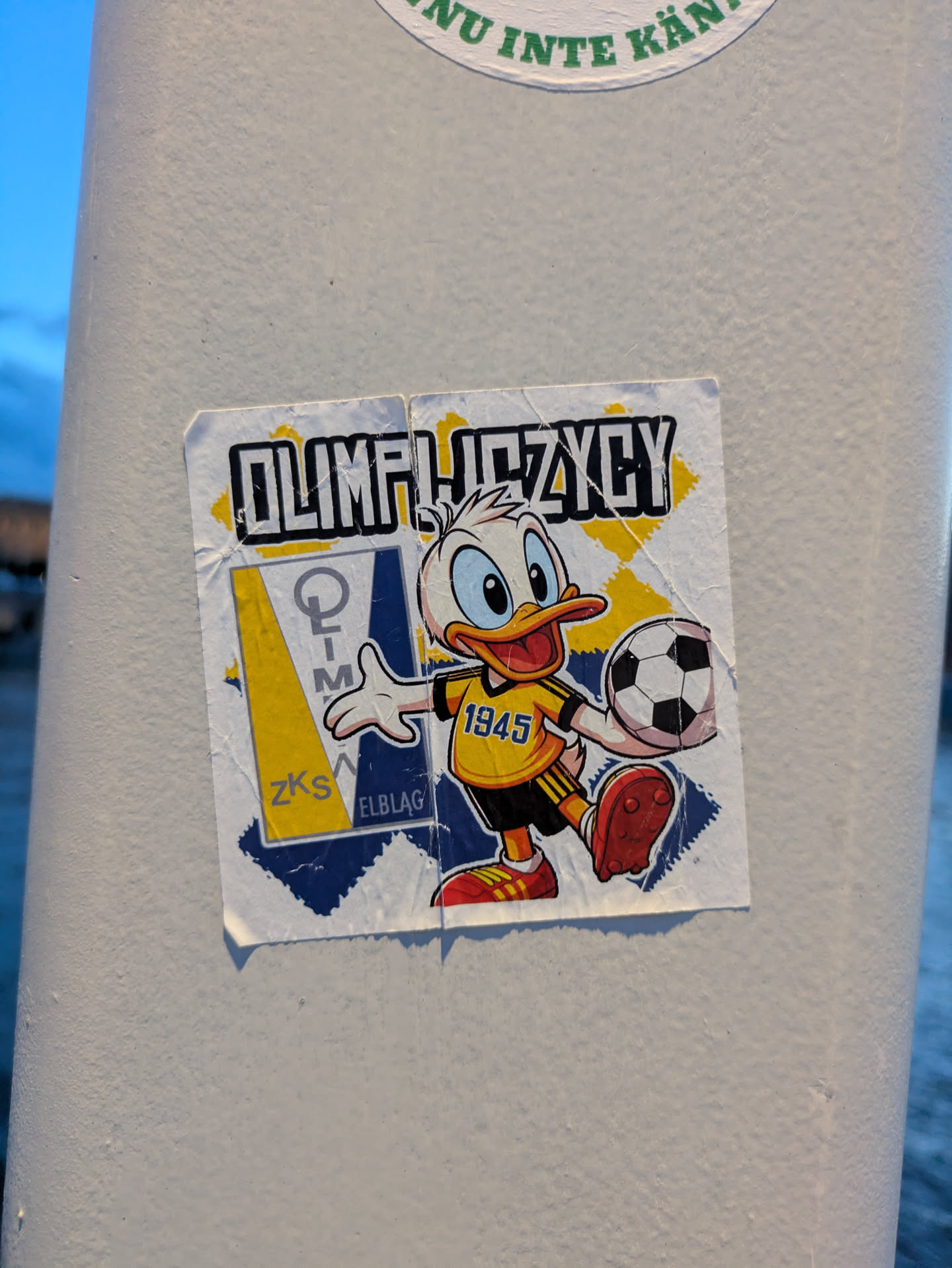 The sticker prominently features a cheerful, cartoon duck character, strongly resembling Disney's Donald Duck, captured in a dynamic pose holding a classic black and white football. The duck is dressed in a vibrant yellow and black striped football jersey with the number '1945' emblazoned on its chest, black shorts, and red cleats with yellow laces. The background of the sticker is divided into a geometric pattern of yellow and deep blue. On the left side, there's a distinct emblem featuring a stylized vertical text element that appears to read 'O-L-I-M', above the letters 'ZKS' and 'ELBLAG'. The top of the sticker displays the word 'OLIMPIJCZYCY' in a bold, outlined font with a yellow fill. The sticker is square-shaped with slightly rounded corners, showing visible signs of wear, including creases and minor fraying along the edges.