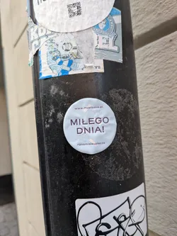 Street sticker A round, light blue sticker featuring the Polish text 'MIŁEGO DNIA!' in purple. It includes the website 'www.mustbake.pl' at the top and the hashtag '#znamisieupiecze' at the bottom, accented by small beige tape graphics.