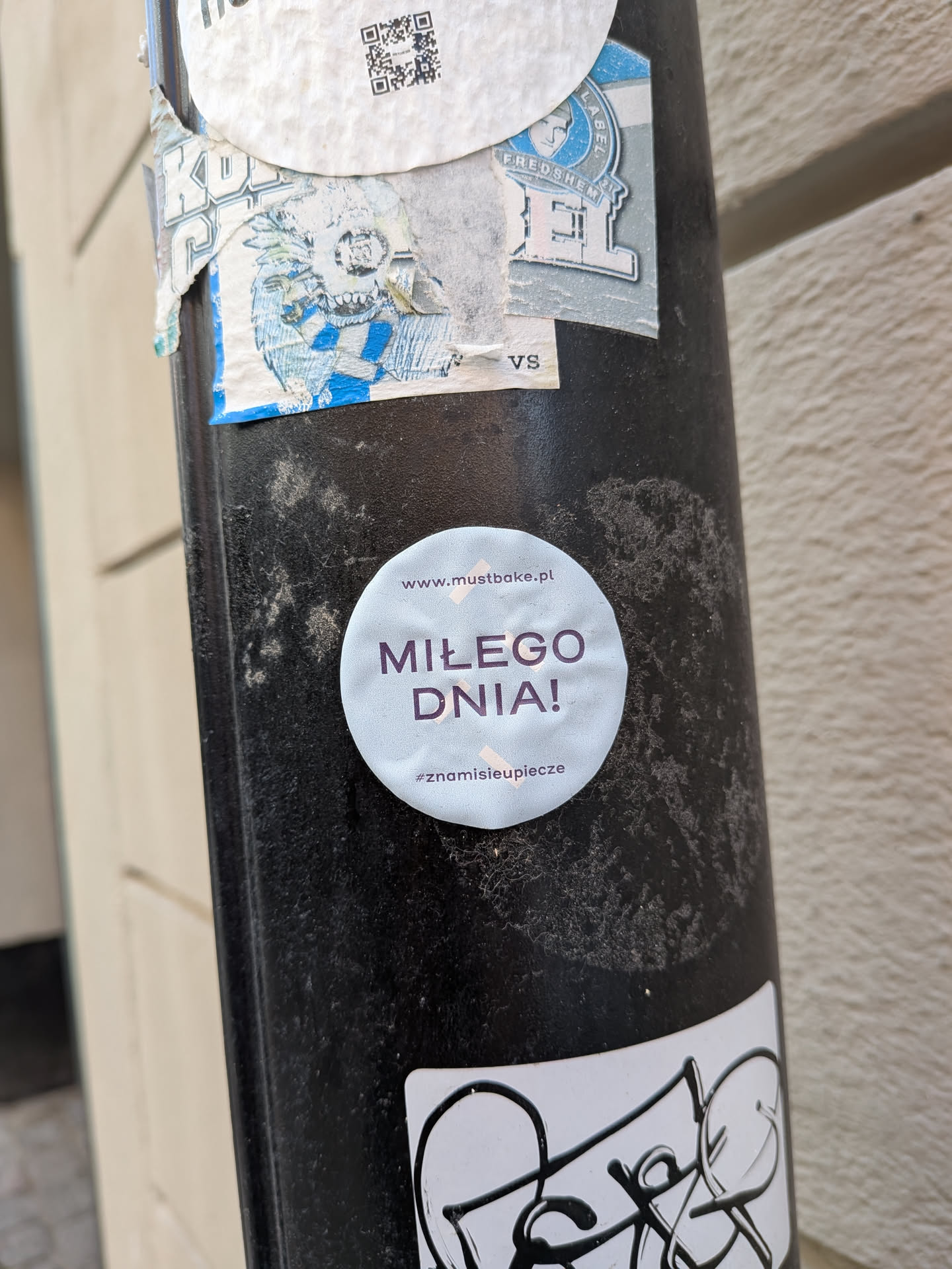 A round, light blue sticker featuring the Polish text 'MIŁEGO DNIA!' in purple. It includes the website 'www.mustbake.pl' at the top and the hashtag '#znamisieupiecze' at the bottom, accented by small beige tape graphics.
