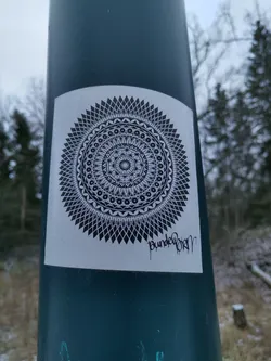Street sticker A square sticker featuring a detailed black and white mandala design. The mandala is surrounded by a geometric pattern, and the sticker is affixed to a teal pole outdoors. The artist's signature, 'Bunderborn', is visible at the bottom.
