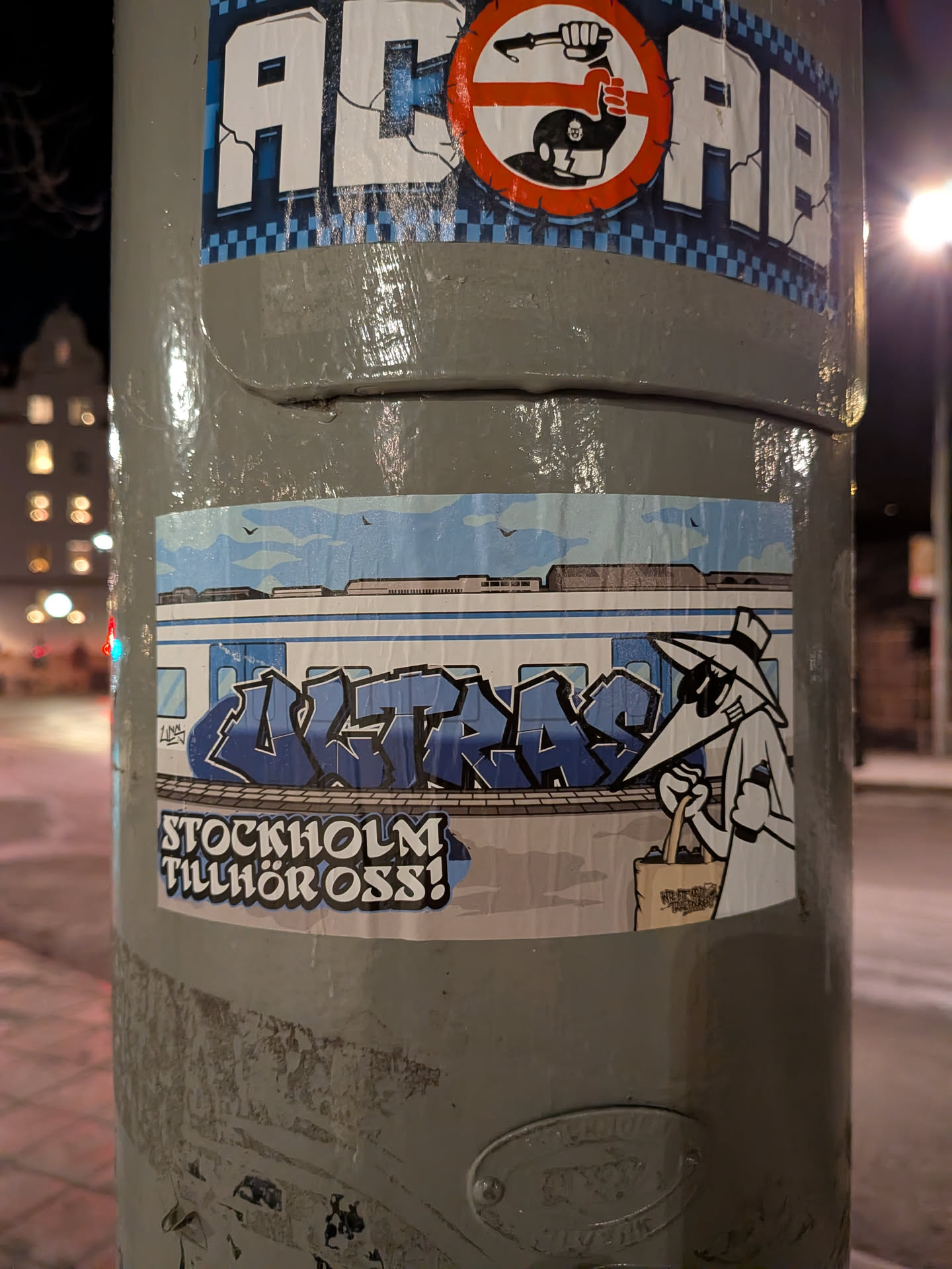 The sticker features a light blue and white train car, prominently tagged with dark blue and light blue bubble-style graffiti spelling 'ULTRAS'. Above the train, a light blue sky with a few small birds and distant city outlines is visible. To the right, a cartoon character, resembling a figure from Spy vs. Spy with a long nose, hat, and sunglasses, stands holding a black spray can in one hand and a light brown tote bag filled with additional spray cans in the other. Below the train and character, the phrase 'STOCKHOLM TILLHÖR OSS!' is written in white letters with a black outline, set against a dark grey, textured background that suggests a platform or ground.