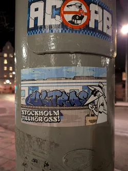Street sticker The sticker features a light blue and white train car, prominently tagged with dark blue and light blue bubble-style graffiti spelling 'ULTRAS'. Above the train, a light blue sky with a few small birds and distant city outlines is visible. To the right, a cartoon character, resembling a figure from Spy vs. Spy with a long nose, hat, and sunglasses, stands holding a black spray can in one hand and a light brown tote bag filled with additional spray cans in the other. Below the train and character, the phrase 'STOCKHOLM TILLHÖR OSS!' is written in white letters with a black outline, set against a dark grey, textured background that suggests a platform or ground.