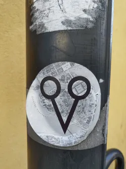 This round sticker features a simple yet striking design. Two circles are connected to a V-shape, creating a unique symbol. The background of the sticker appears to be a grayscale map, adding an interesting juxtaposition of modern design and historical context. The sticker is attached to a dark-colored pole in an outdoor setting.