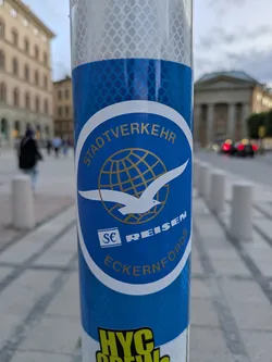 Street sticker The sticker is predominantly blue and white with gold lettering. It features a circular emblem at its center. Within the circle, a stylized white seagull is depicted in flight, with its wings spread. Behind the seagull is a golden globe with a grid pattern representing longitude and latitude lines. The upper arc of the circle contains the text 'STADTVERKEHR' in gold capital letters. Below the seagull and globe, the words 'REISEN' and 'ECKERÜTFÖRDE' are also present in gold, with 'REISEN' on a white background and 'ECKERÜTFÖRDE' on a blue background. The entire sticker is affixed to a reflective, diamond-patterned surface, likely a pole.