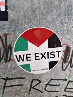 Street sticker Round sticker with a color block design resembling a flag and the text "WE EXIST" in black letters.  The website www.weexist.info is printed at the bottom. The sticker is affixed to a textured gray surface.