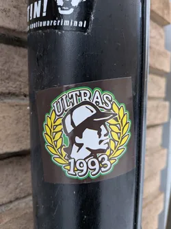 Street sticker The sticker is a rectangular, dark brown decal featuring a central circular emblem. The emblem has a vibrant green outer border. At the top, the word "ULTRAS" is written in white capital letters with a green outline, following the curve of the circle. The center of the emblem displays a stylized profile of a person's head wearing a baseball cap, facing towards the right. This head is rendered in white with black outlines and shading. Flanking the head on both sides is a yellow laurel wreath, also outlined in green, symbolizing victory or honor. At the bottom of the emblem, the year "1993" is displayed in white numbers with a green outline, mirroring the curve of the circle.