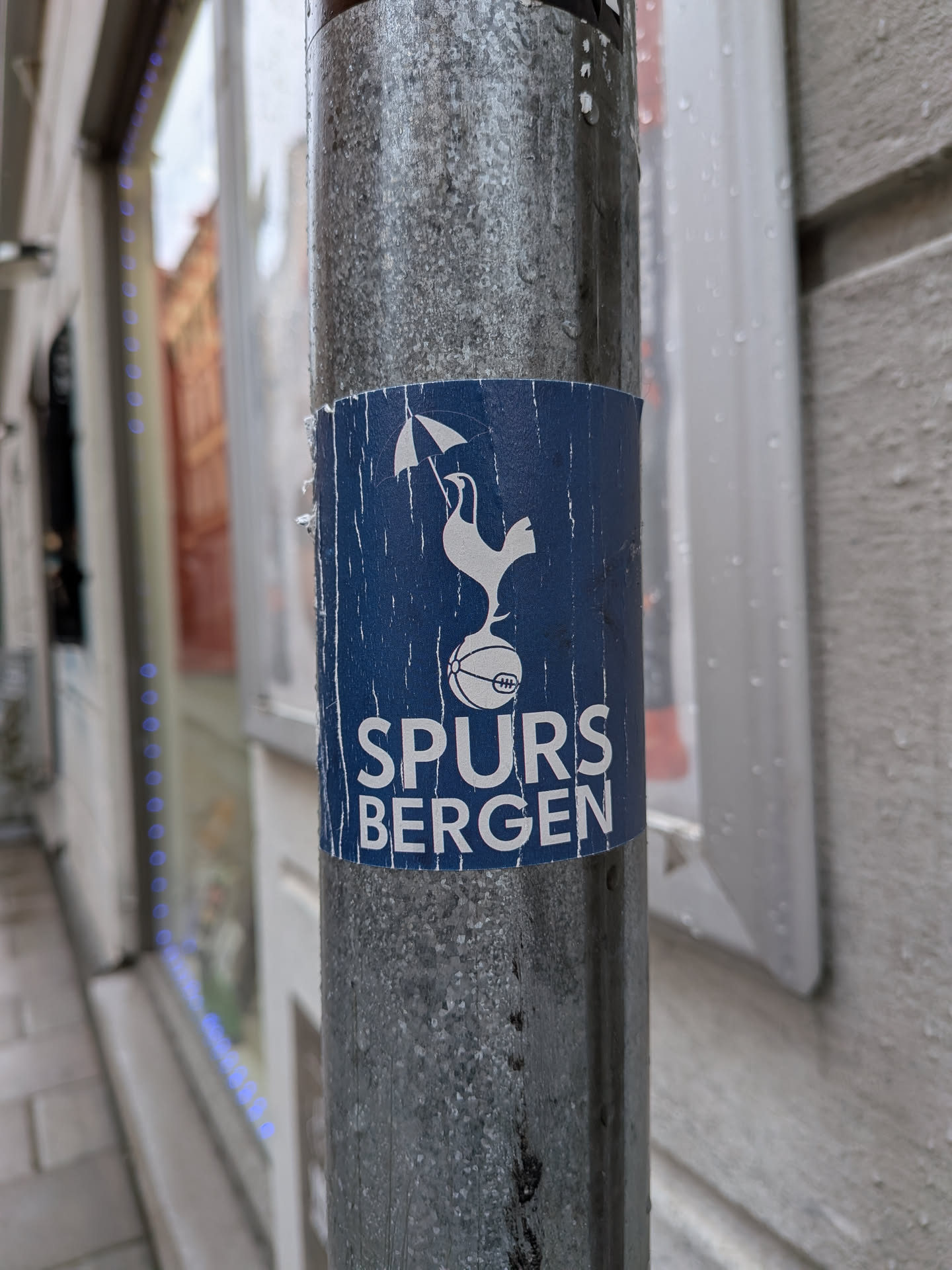 A rectangular sticker with a dark blue background, affixed to a textured metal pole. The sticker features a white illustration of the Tottenham Hotspur cockerel logo, but with distinct modifications. Above the cockerel, a white umbrella with black stripes is depicted. The cockerel itself stands on a white rugby ball, replacing the traditional football. Below the graphic, the words "SPURS" and "BERGEN" are stacked in bold, white capital letters. The sticker shows signs of wear, with vertical white streaks and slightly frayed edges, and is covered in small water droplets, matching the wet appearance of the pole.