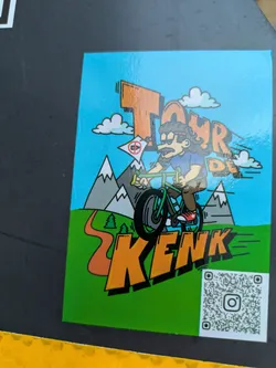 The sticker features a vibrant cartoon illustration of a person riding a bicycle with large wheels, possibly a BMX or mountain bike. The rider has brown curly hair, sunglasses, a mustache, and is wearing a blue t-shirt and brown shorts. The background is a bright sky blue, with stylized white clouds. In the middle ground, there are grey mountains with snow-capped peaks and a winding orange path leading up to them. The foreground is a bright green grassy area. Prominently displayed in large, orange, blocky letters outlined in black is the text 'KENK'. In the bottom right corner, there is a QR code with an Instagram logo in the center.