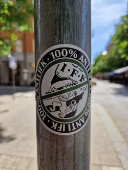 Street sticker A round sticker with the text "100% Anti IFK" written in a circle.  The inner circle features a graphic of a lion emblem crossed out. The sticker is affixed to a metal pole.