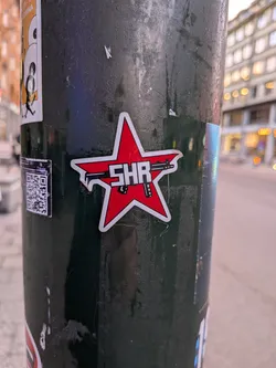 The sticker is a red star with a white outline. Inside the star, a black silhouette of a rifle is depicted horizontally, with its barrel pointing to the left and the stock to the right. The letters 'SHR' are prominently displayed in white, bold, sans-serif font across the center of the star, overlapping the rifle. The overall design is graphic and impactful.