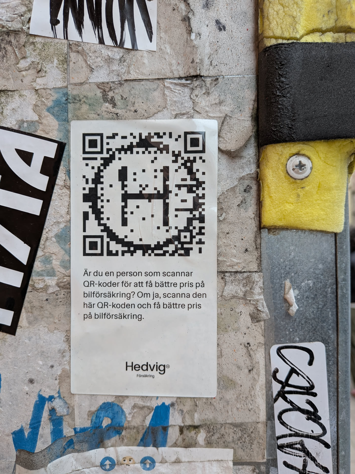 A rectangular white sticker pasted on a textured wall. The top half of the sticker features a large black QR code with a stylized letter 'H' integrated into its center. Below the QR code, there is a short paragraph of Swedish text, and at the very bottom, the brand logo 'Hedvig® Försäkring' is printed in a clean, sans-serif font.