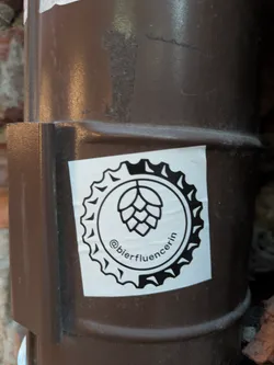 Street sticker The sticker is circular with a white background. It features a black outline of a hop cone in the center, surrounded by a circular border designed to resemble the jagged edge of a bottle cap. Below the hop cone, the text "@bierfluencerin" is written in a black, cursive font, following the curve of the circle.