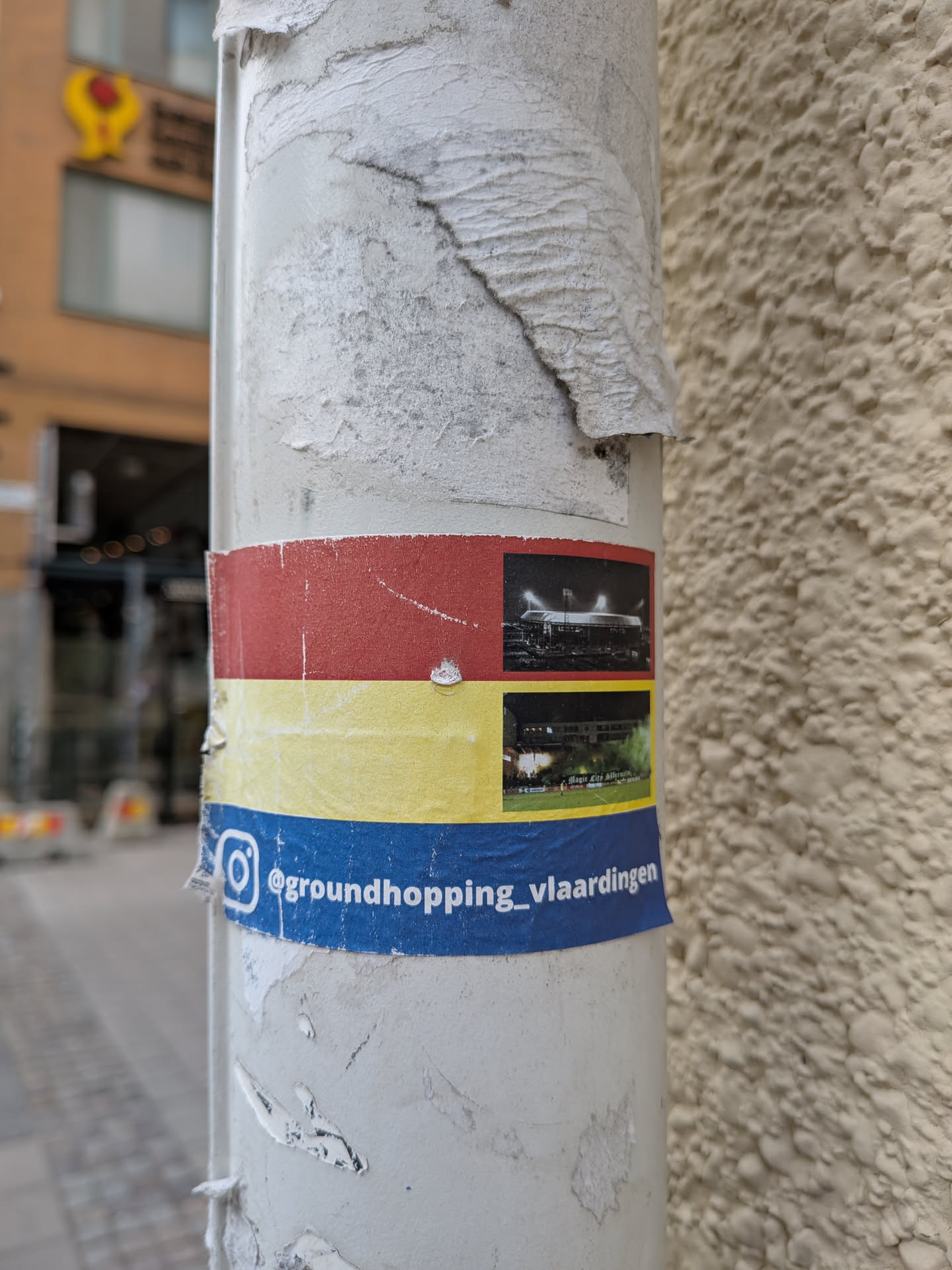 Street sticker The sticker is horizontally oriented and consists of three colored bands: red at the top, yellow in the middle, and blue at the bottom. The red and yellow bands each contain a photograph of a football stadium. The top photograph shows a stadium at night with illuminated stands. The bottom photograph also depicts a stadium at night, with green smoke or flares visible on the field, and text that appears to read 'Magisch Elf Vlaardingen'. The blue band prominently features an Instagram logo (a stylized camera icon) and the text '@groundhopping_vlaardingen'. The sticker exhibits some wear and tear, with slight peeling at the edges.