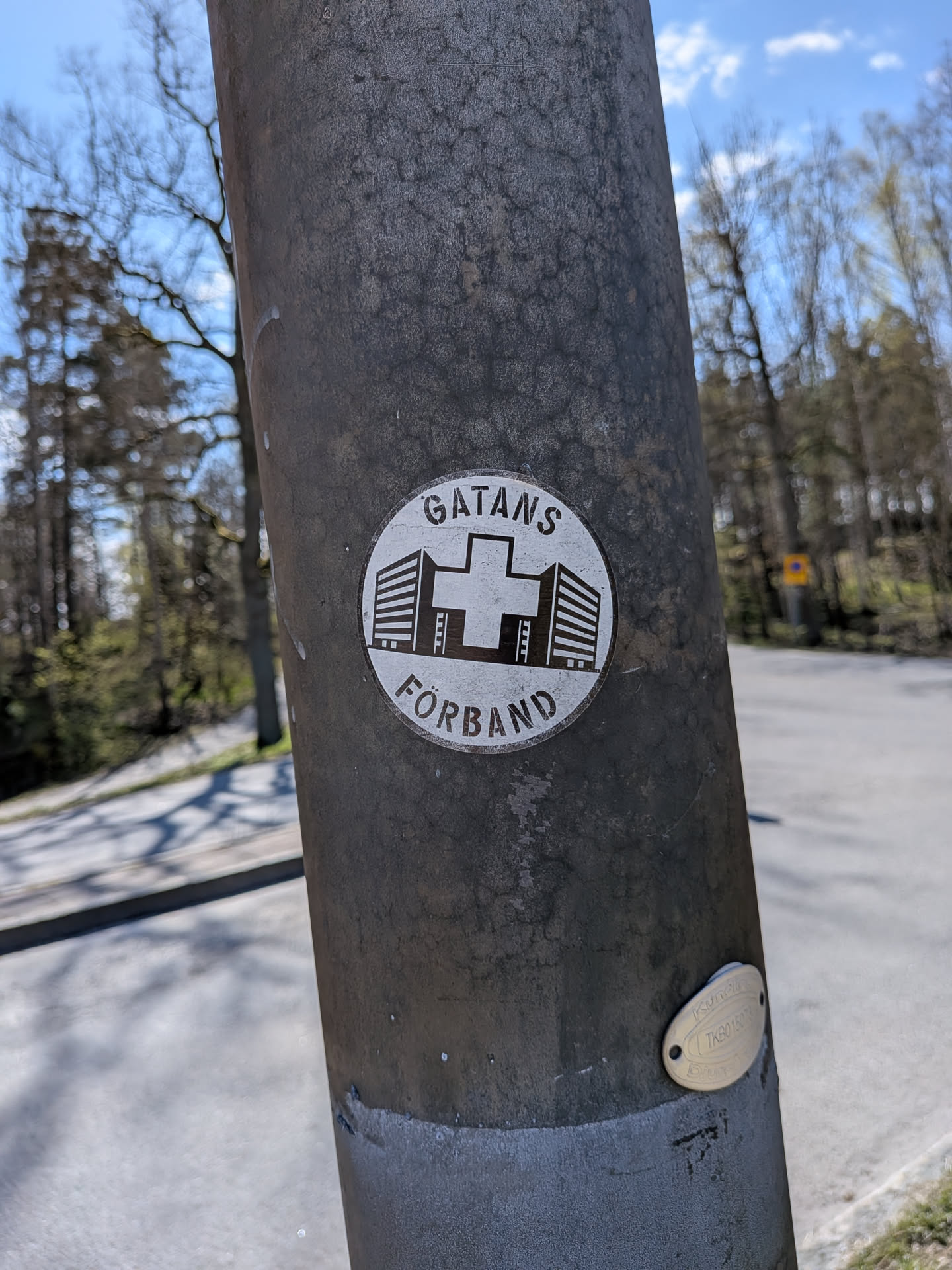 A circular sticker featuring a monochrome design with a white background and dark (likely black or dark grey) elements. In the center, a prominent cross symbol is flanked by stylized illustrations of multi-story buildings on both its left and right sides, depicted with horizontal lines representing windows. The text "GATANS" is arched along the top edge of the circle, and "FÖRBRAND" is arched along the bottom edge, both in capital letters. The sticker is adhered to a textured, dark metal pole and shows some signs of wear.